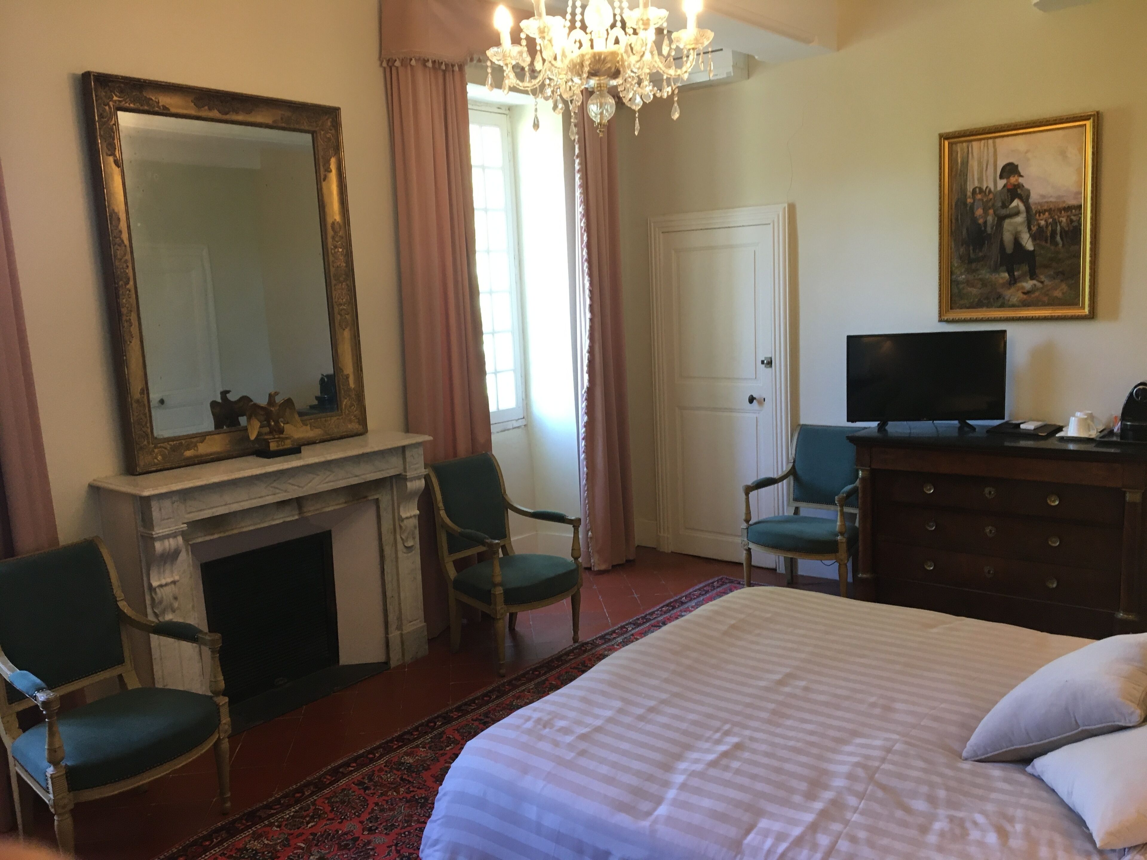 A hotel room with a large bed, two chairs, a television, a fireplace, a painting, and a chandelier.