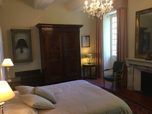 Un dormitorio con una cama grande, un armario de madera, una araña, una mesita con una lámpara, una silla y una chimenea.