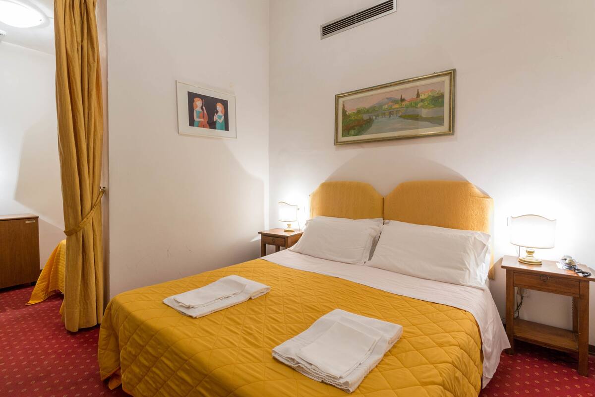 Ein Hotelzimmer mit einem Bett, zwei Nachttischen, einem Schrank und einem Bild an der Wand sowie einem Fenster mit Vorhängen.