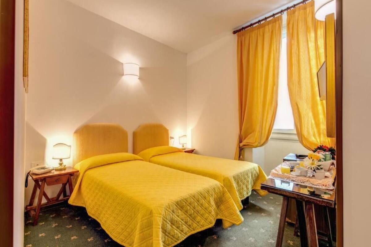 Ein Zimmer mit zwei Betten, einem Nachttisch mit Lampe, einem kleinen Tisch mit Frühstücksgebäck und gelben Vorhängen.
