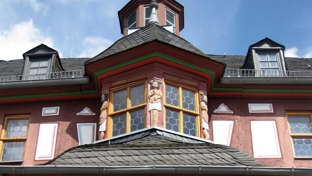 Un edificio con una columna central destacada, un ventanal saliente y una pequeña torre con veleta.