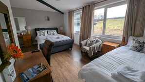 A hotel room with a large bed, a smaller bed, a desk with a lamp, a chair, and a window with a view of the countryside. - Old North Inn Hotel (Inverness)