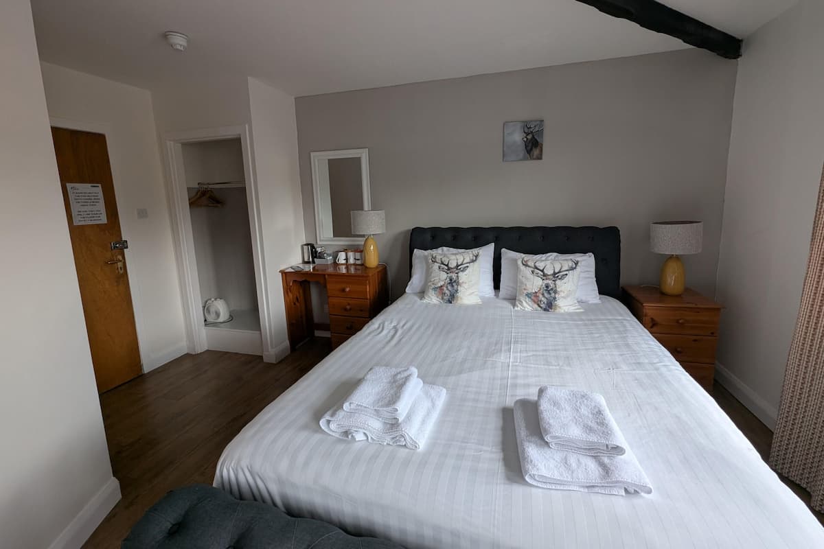 A neatly made bed with white linens and decorative pillows, a bedside table with a lamp, a wooden desk with a chair, and a coat rack.