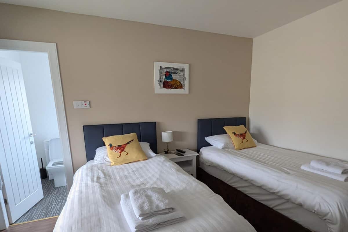 A hotel room with two beds, each with white linens and a single pillow. There is a small table between the beds with a lamp and a remote control. A painting hangs on the wall above the beds.