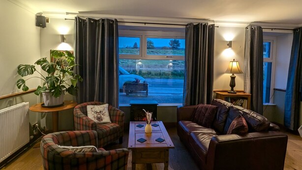 A cozy living room with a fireplace, two plaid armchairs, a sofa, and a view of a parking lot through large windows. - Old North Inn Hotel (Inverness)