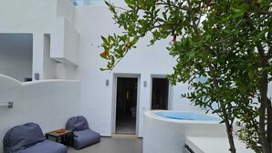 Un espace extérieur moderne avec un mur blanc incurvé, un petit coin salon avec deux fauteuils en forme de poire, une petite table avec un panier, et un arbre aux feuilles vertes et aux fleurs rouges.