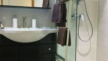 Een badkamer met een douchecabine van glas, een wastafel met spiegel erboven en handdoeken aan de douchedeur.