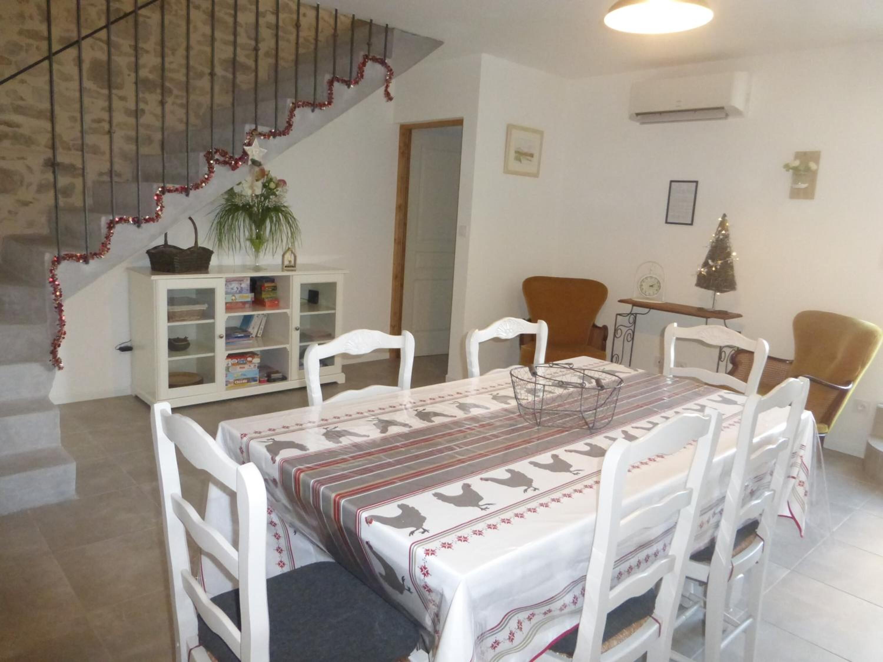 Un comedor con una mesa dispuesta para seis personas, sillas blancas, un armario blanco con libros, una planta pequeña y una escalera adornada con guirnaldas rojas.