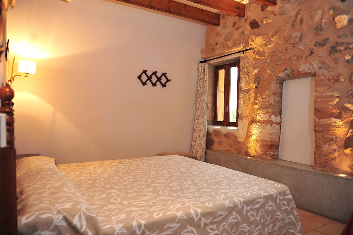 A bedroom with a bed, a wall-mounted light, a window with curtains, and a stone wall.
