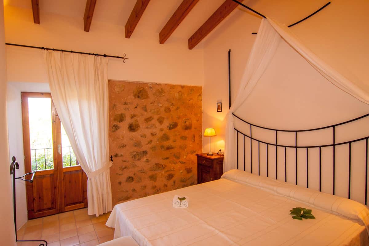 A bedroom with a stone wall, a bed with a canopy, a nightstand, and a window with curtains.