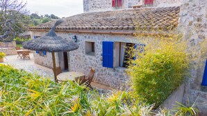 A stone building with a tiled roof, a thatched roof structure, and a small table with chairs outside. - Finca Ses Cases Noves (Sant Llorenc des Cardassar)
