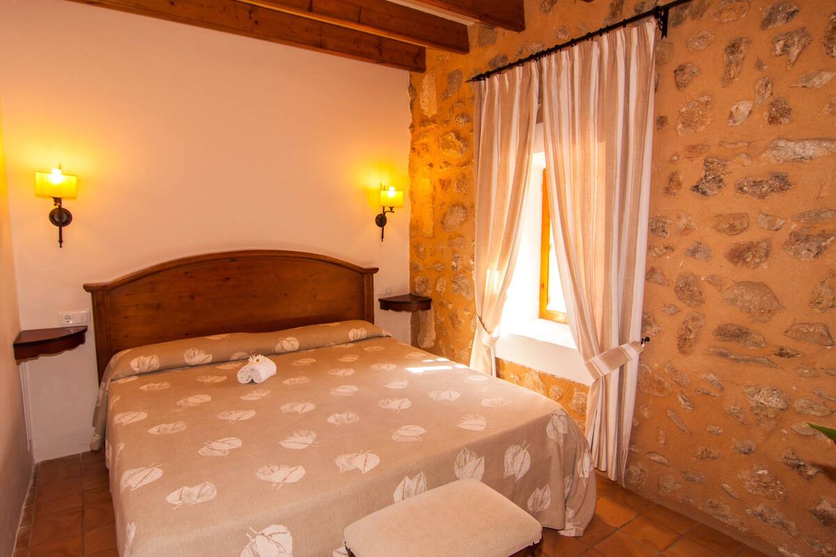 A bedroom with a wooden headboard, two bedside lamps, a bed with a leaf-patterned bedspread, a bench, and a stone-patterned wall.