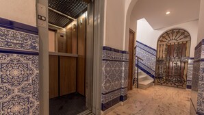 Un ascensor de diseño moderno, un pasillo con piso de mármol y baldosas azules y blancas con diseño, y una escalera con una puerta metálica decorativa.