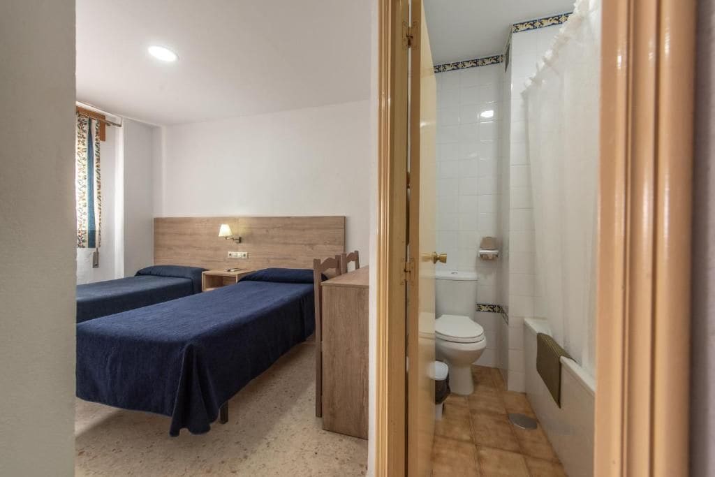 A hotel room with two beds, a wooden headboard, a bedside table with a lamp, a small wooden dresser, and a bathroom with a toilet and shower.