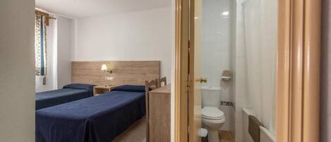 A hotel room with two beds, a wooden headboard, a bedside table with a lamp, a small wooden dresser, and a bathroom with a toilet and shower.
