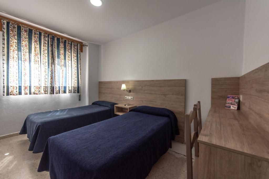 Een kamer met twee eenpersoonsbedden, een houten hoofdbord, een nachtkastje met een lamp en een raam met patroon gordijnen.