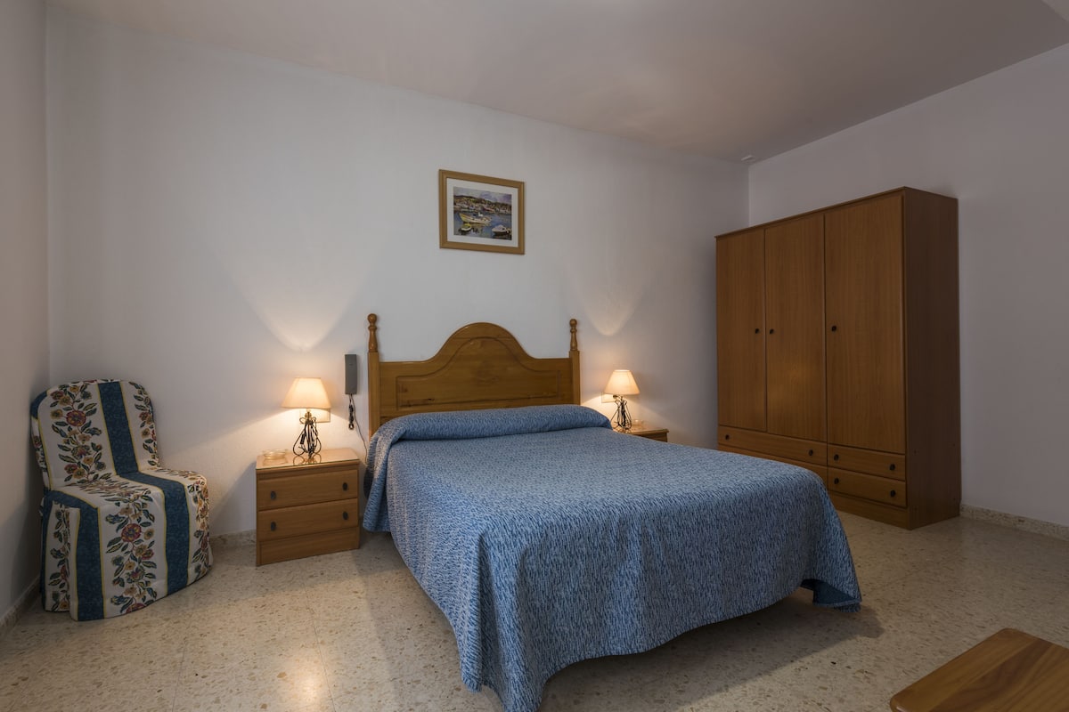 Een slaapkamer met een bed, nachtkastjes, een houten garderobe, een stoel en een aan de muur bevestigd schilderij.