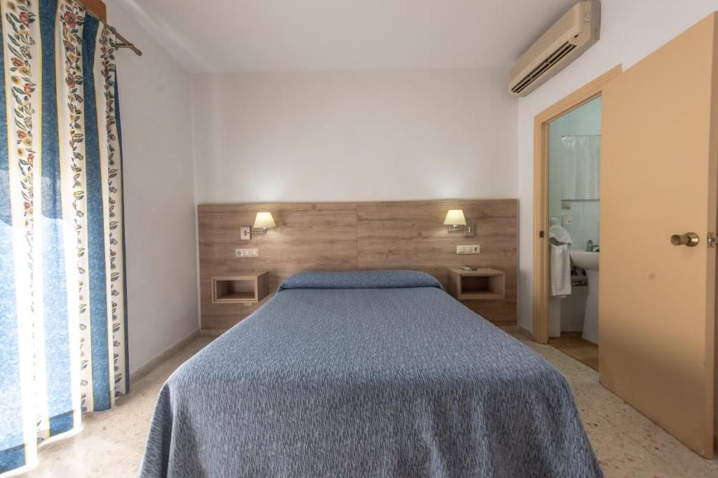 Ein Schlafzimmer mit einem hölzernen Kopfteil, zwei Nachttischlampen, einem Bett mit blauer Bettdecke und einem durchgehenden Spalt zu einem Badezimmer. Ein Vorhang mit einem Blumenmuster ist ebenfalls zu sehen.