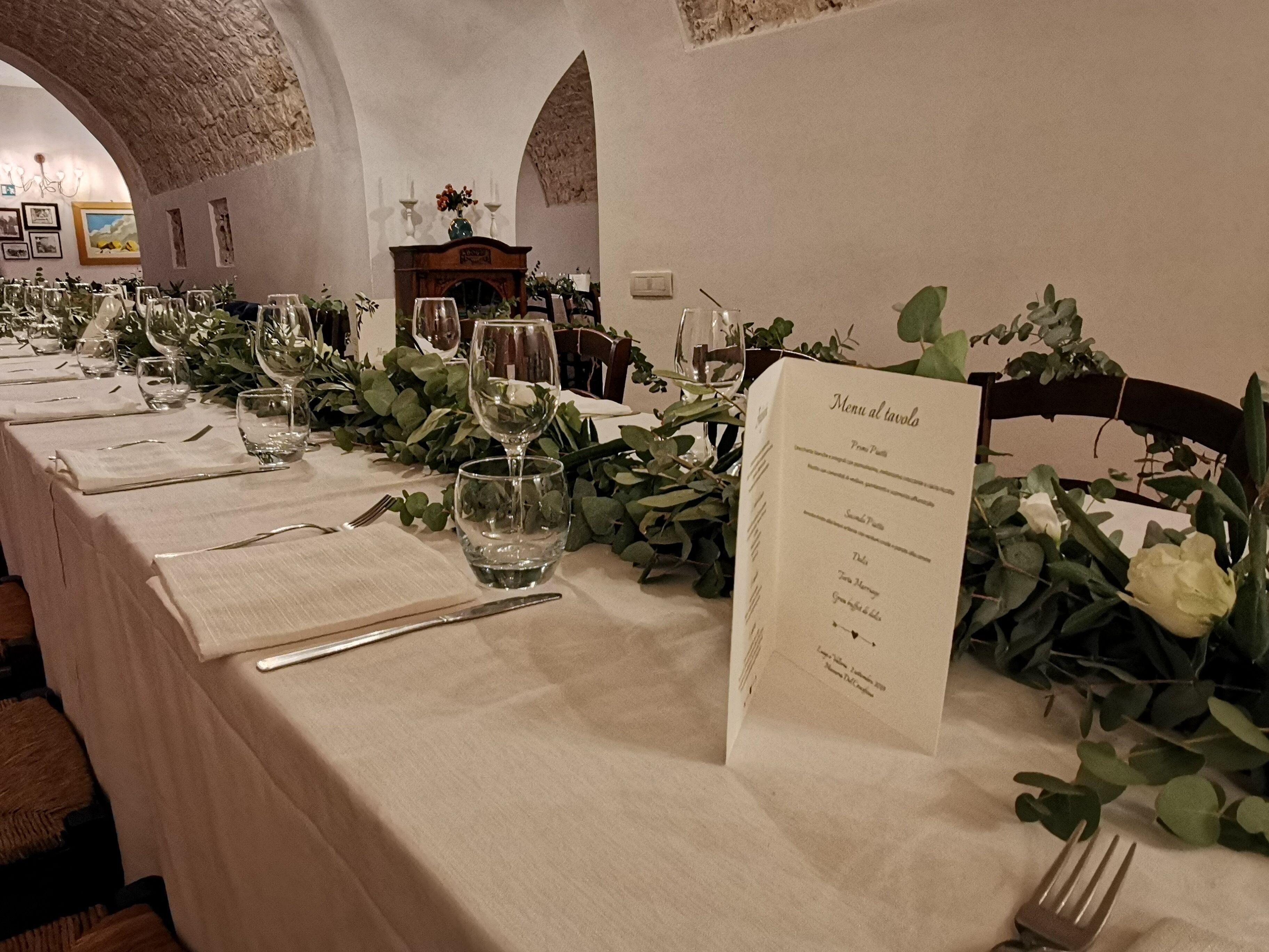 A long dining table set with wine glasses, cutlery, and menus, surrounded by greenery and a fireplace.