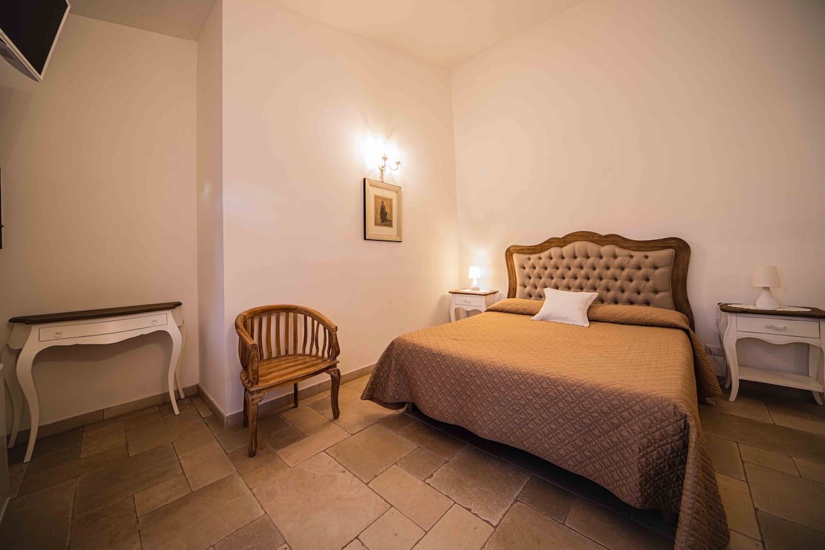 A bedroom with a large bed, a wooden chair, a bedside table, and a picture on the wall.