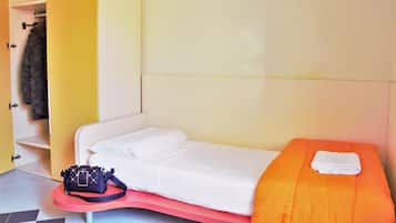 Una cama individual con ropa de cama blanca y una manta naranja, una mesita con un bolso negro y un armario amarillo con un abrigo colgado.