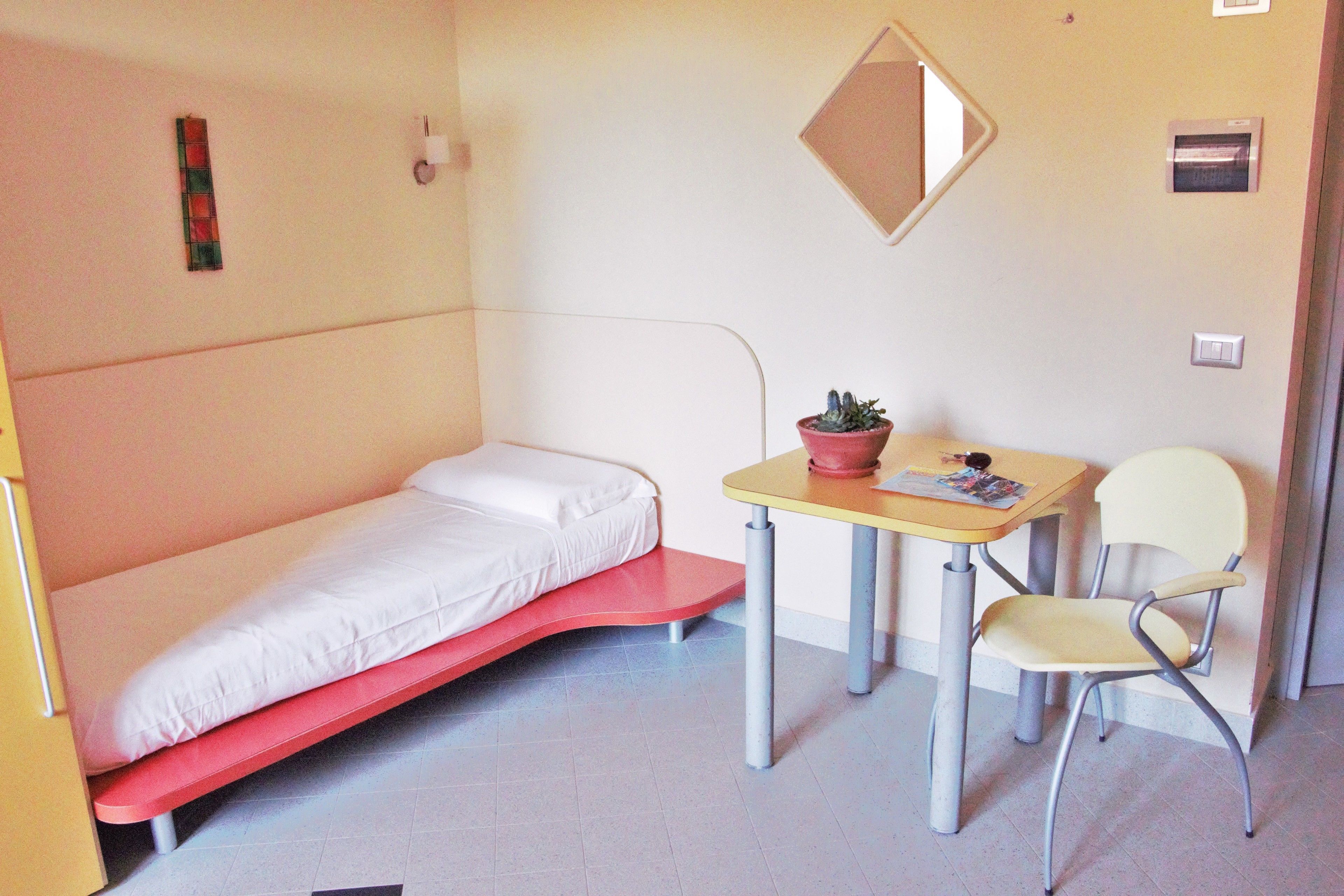 Een kleine, compacte kamer met een enkel bed, een tafeltje met stoel en een potplant.