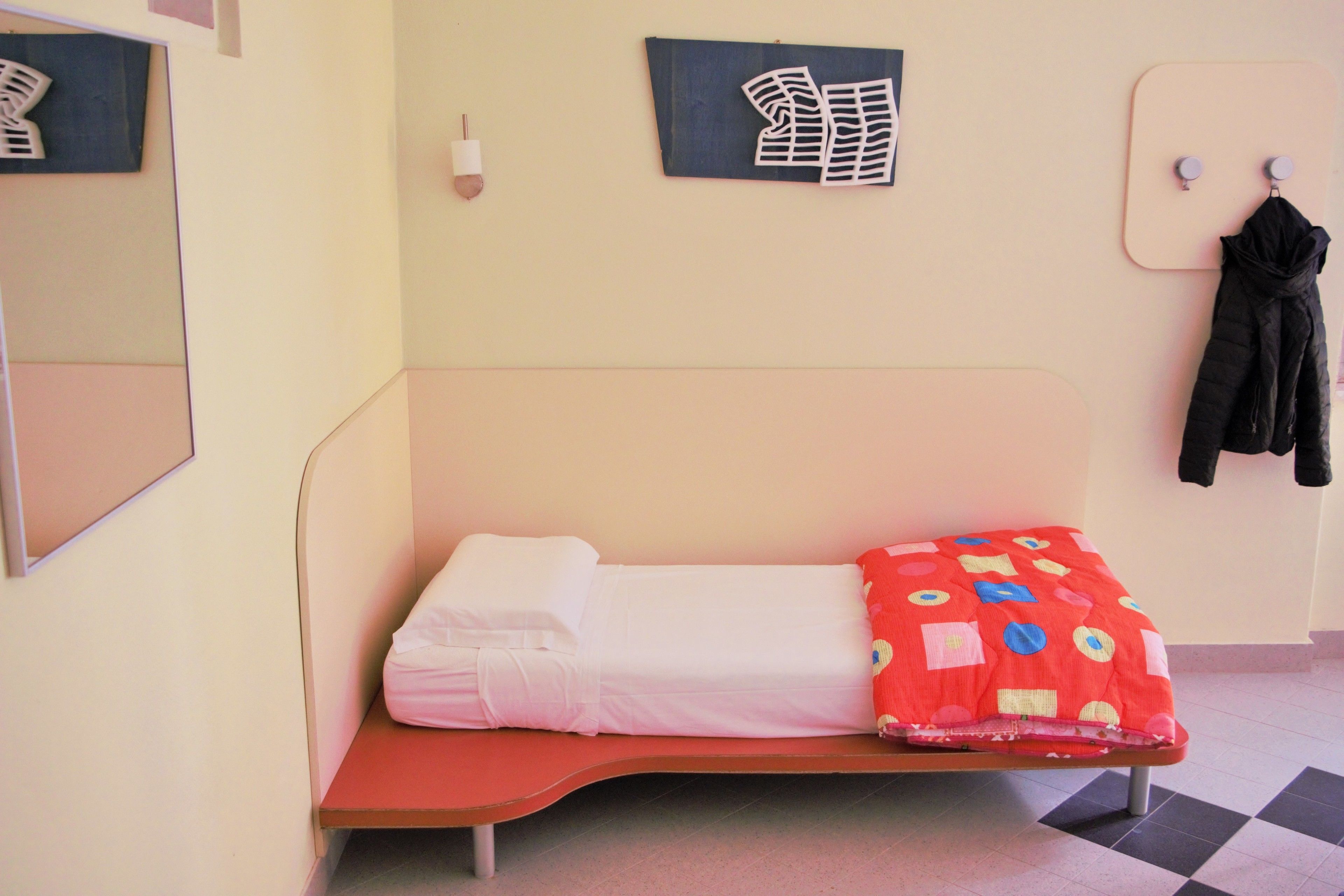 Ein Einzelbett mit roter Decke, einem weißen Kopfkissen und einem schwarzen Jacket an einem Haken.