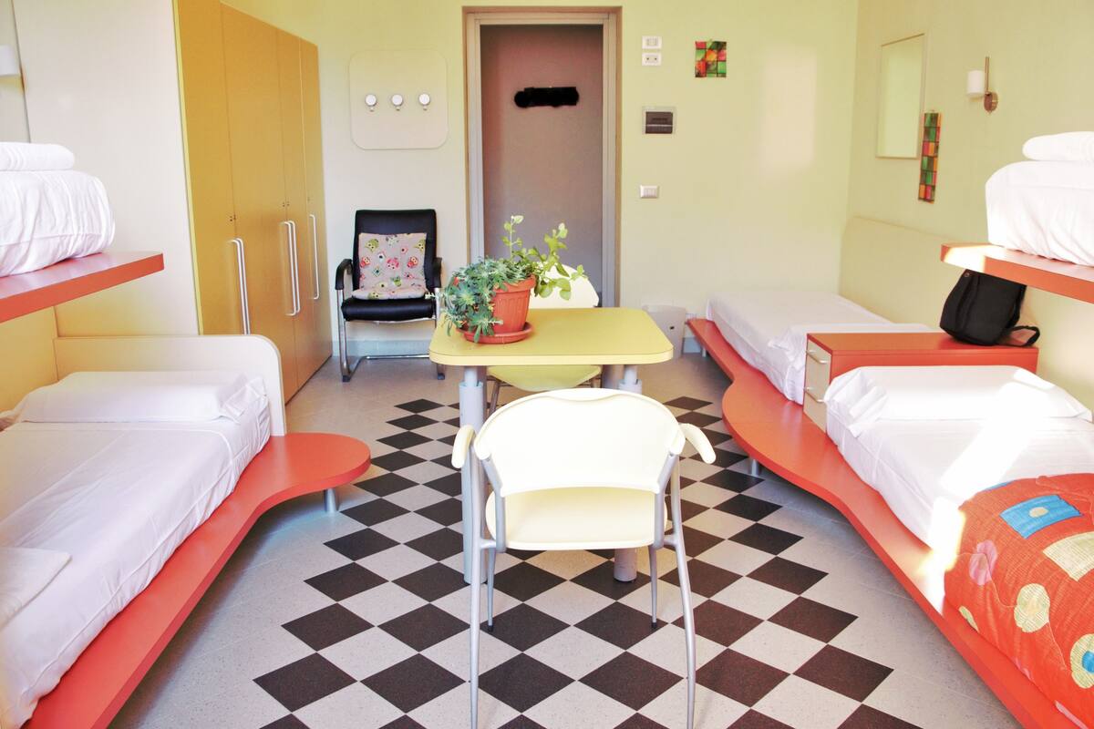 Hotellihuone, jossa on kerrossänky, yksi erillinen sänky, pieni pöytä, jossa on kasvi, tuoli ja ruutukuviollinen lattia.