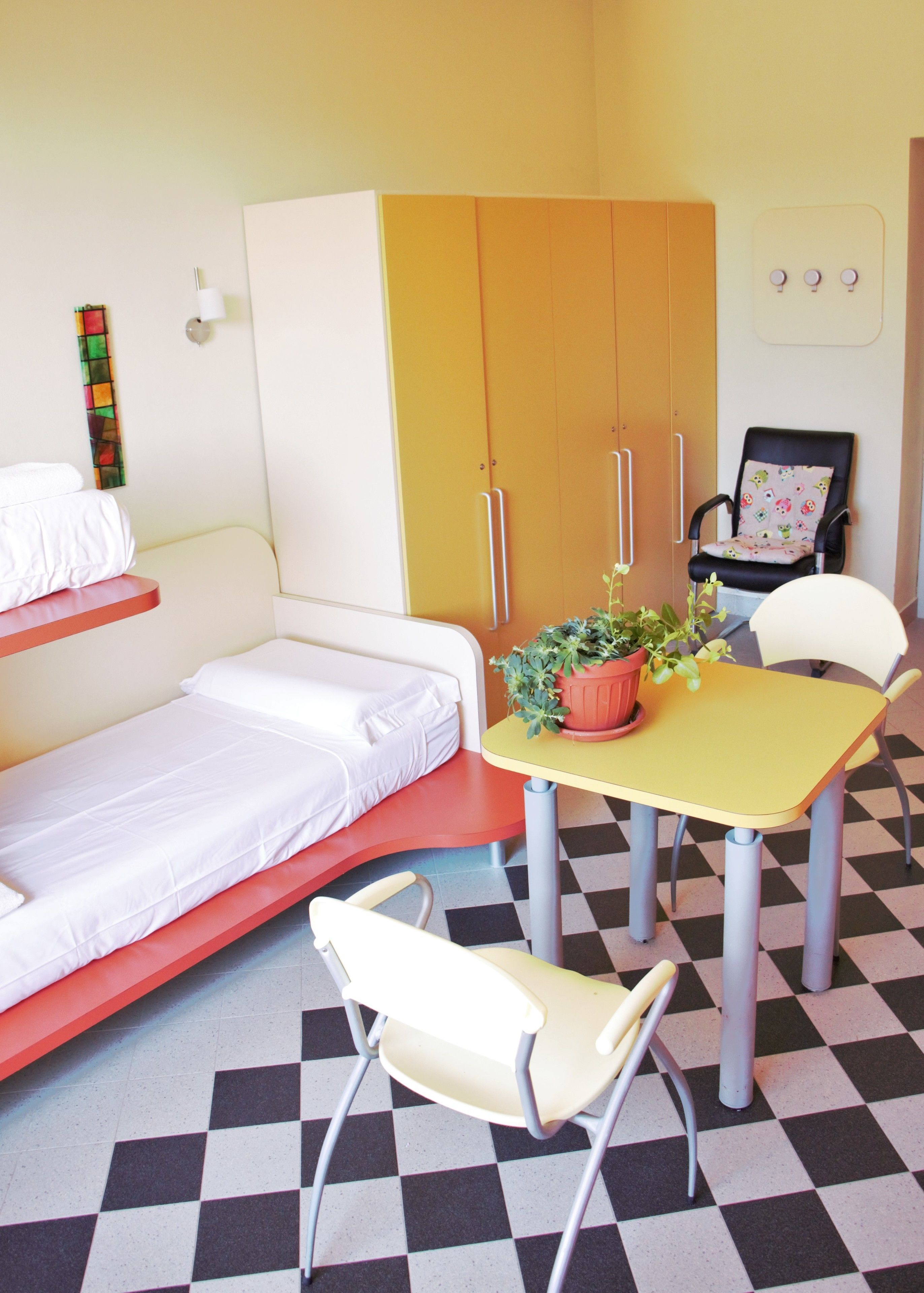 Una habitación pequeña e iluminada, con una cama, una mesa con una planta en maceta, una silla y un armario.