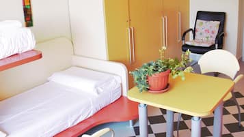 Una habitación pequeña e iluminada, con una cama, una mesa con una planta en maceta, una silla y un armario.
