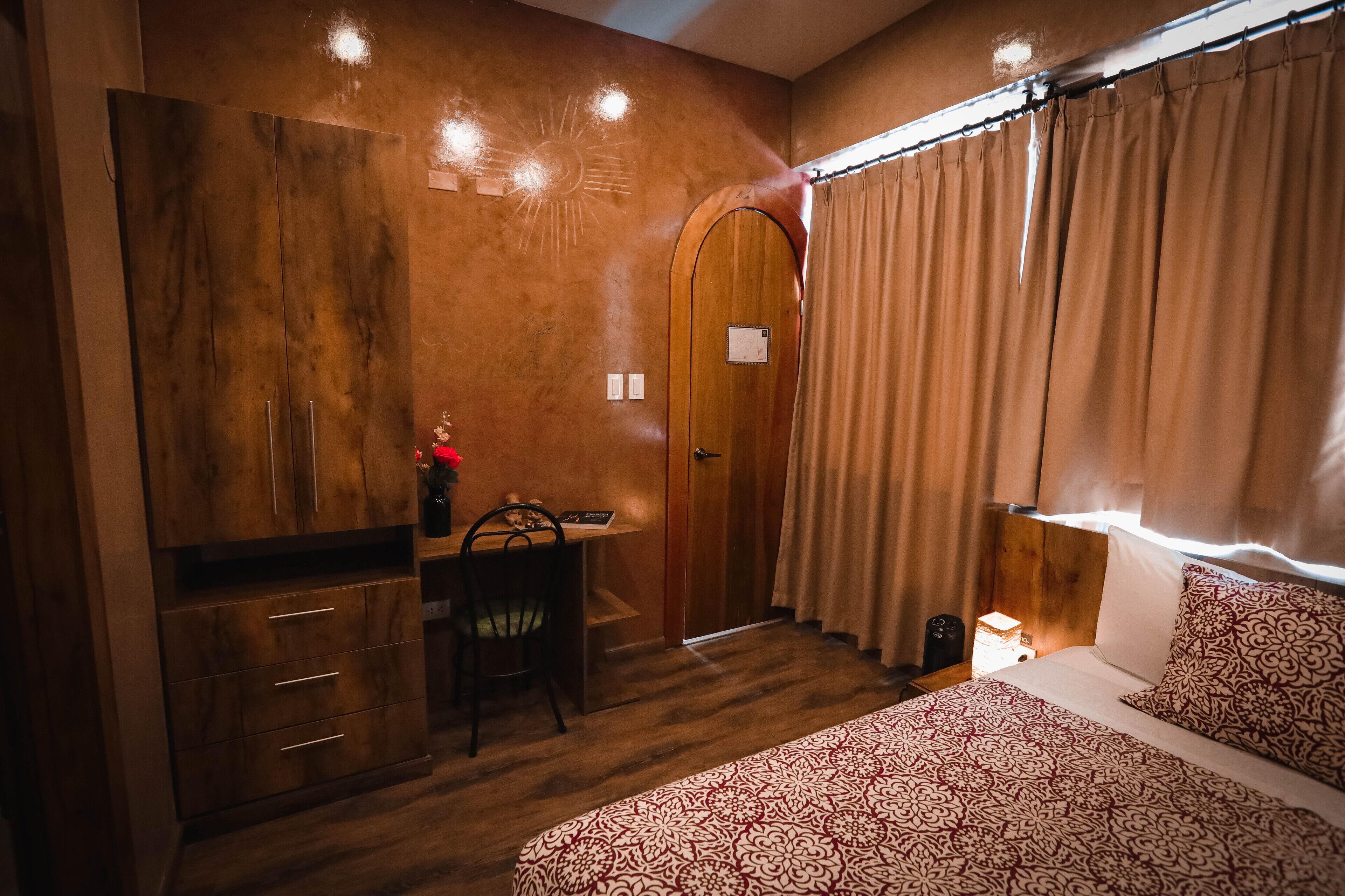 Un dormitorio con un armario de madera, un escritorio con una silla, una cama con ropa de cama estampada y una ventana con cortinas.