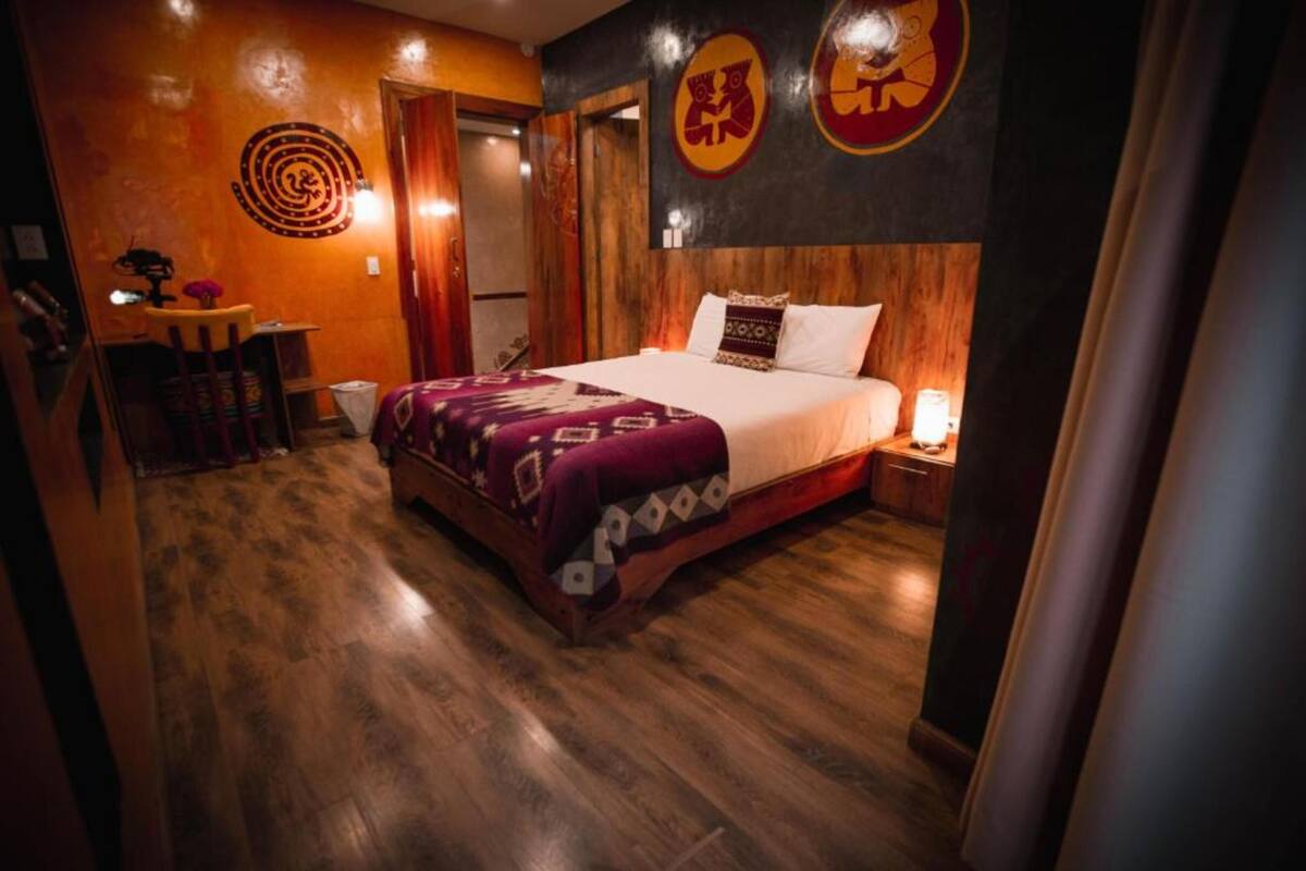 A bedroom with a wooden floor, a bed with a patterned blanket, a nightstand with a lamp, and a wall with circular designs.