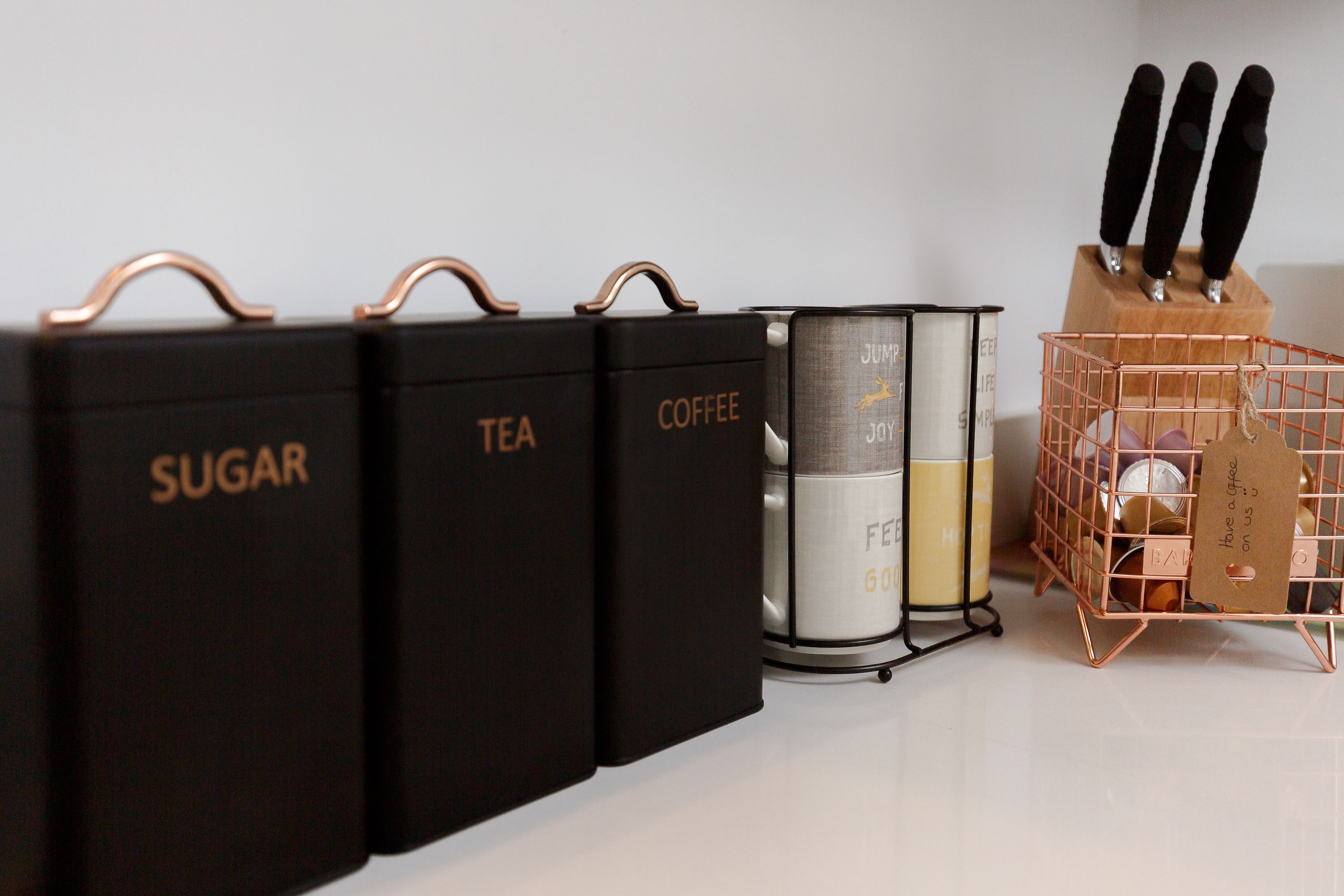 Tres contenedores negros para almacenamiento con etiquetas que dicen 'AZÚCAR', 'TÉ' y 'CAFÉ', un recipiente metálico y una cesta de alambre de cobre con una etiqueta.