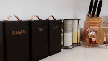 Tres contenedores negros para almacenamiento con etiquetas que dicen 'AZÚCAR', 'TÉ' y 'CAFÉ', un recipiente metálico y una cesta de alambre de cobre con una etiqueta.