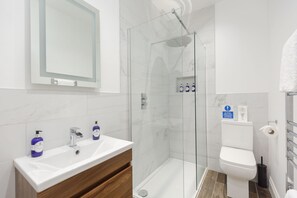 Ein modernes Badezimmer mit einer gläsernen Duschkabine, einem weißen Waschbecken mit Holzschrank und einer Toilette mit Papierhandtuchspender.