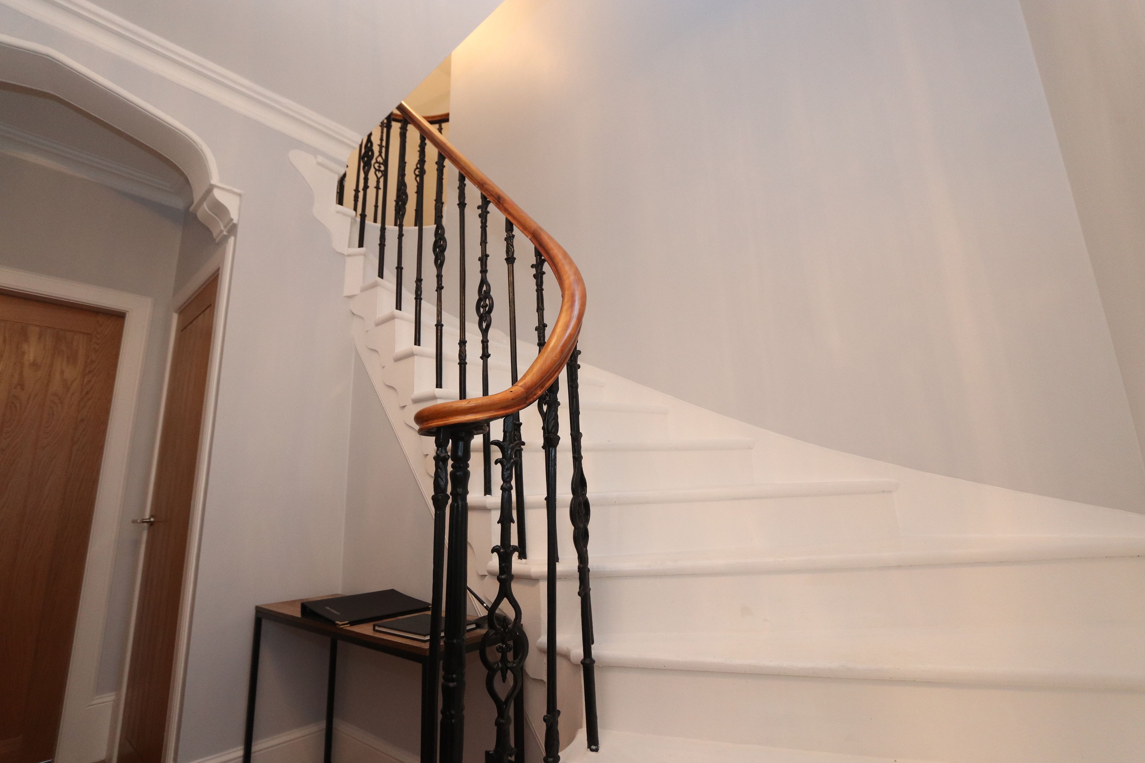 A staircase with a wooden handrail and black balusters, leading to a landing with a white wall and a wooden door.
