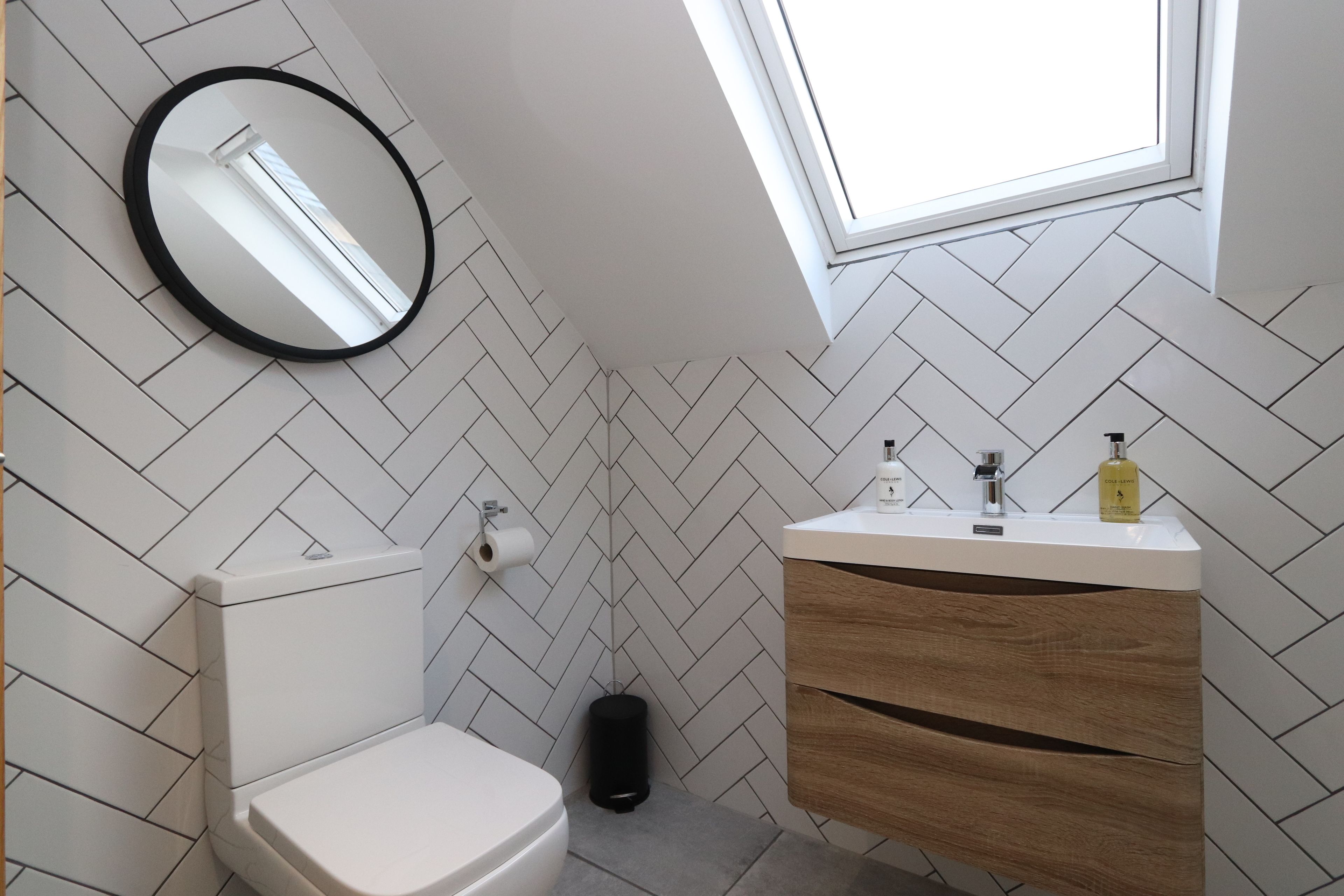 A modern bathroom with a white herringbone tile pattern, a round mirror, a toilet, a sink with a wooden vanity, and a skylight.