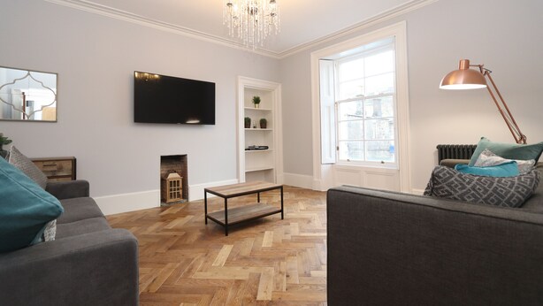 A living room with a herringbone wood floor, a grey sofa, a television mounted on the wall, a fireplace, and a window with a view. - Dollar Apartments at 41 (Dollar)