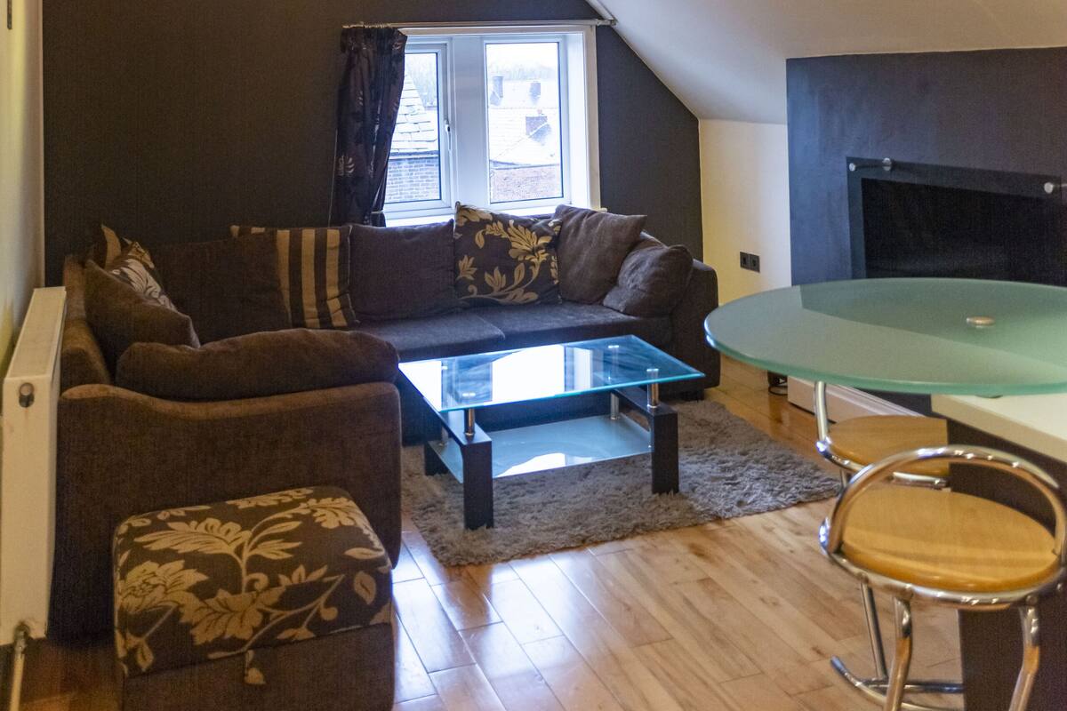 Un salon chaleureux avec un canapé, un pouf et une table basse en verre. On y trouve une cheminée et une fenêtre avec des rideaux.