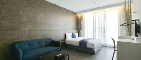 Et moderne hotelværelse med sofa, et lille rundt bord, en seng med et hvidt og lilla tæppe samt et skrivebord med et spejl.