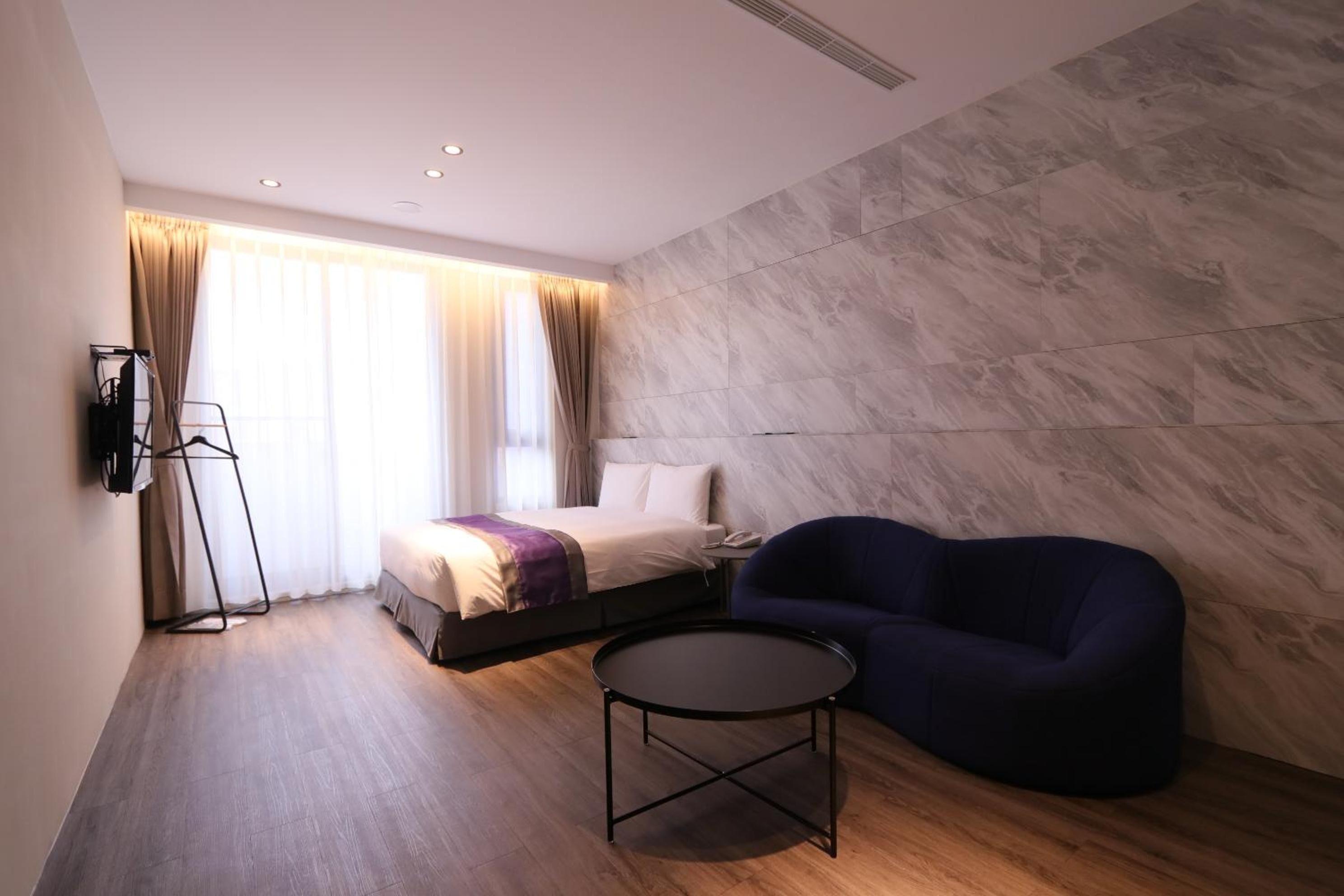 A modern hotel room with a bed, a blue sofa, a small round table, and a large window with curtains.