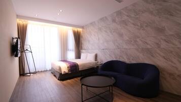 A modern hotel room with a bed, a blue sofa, a small round table, and a large window with curtains.