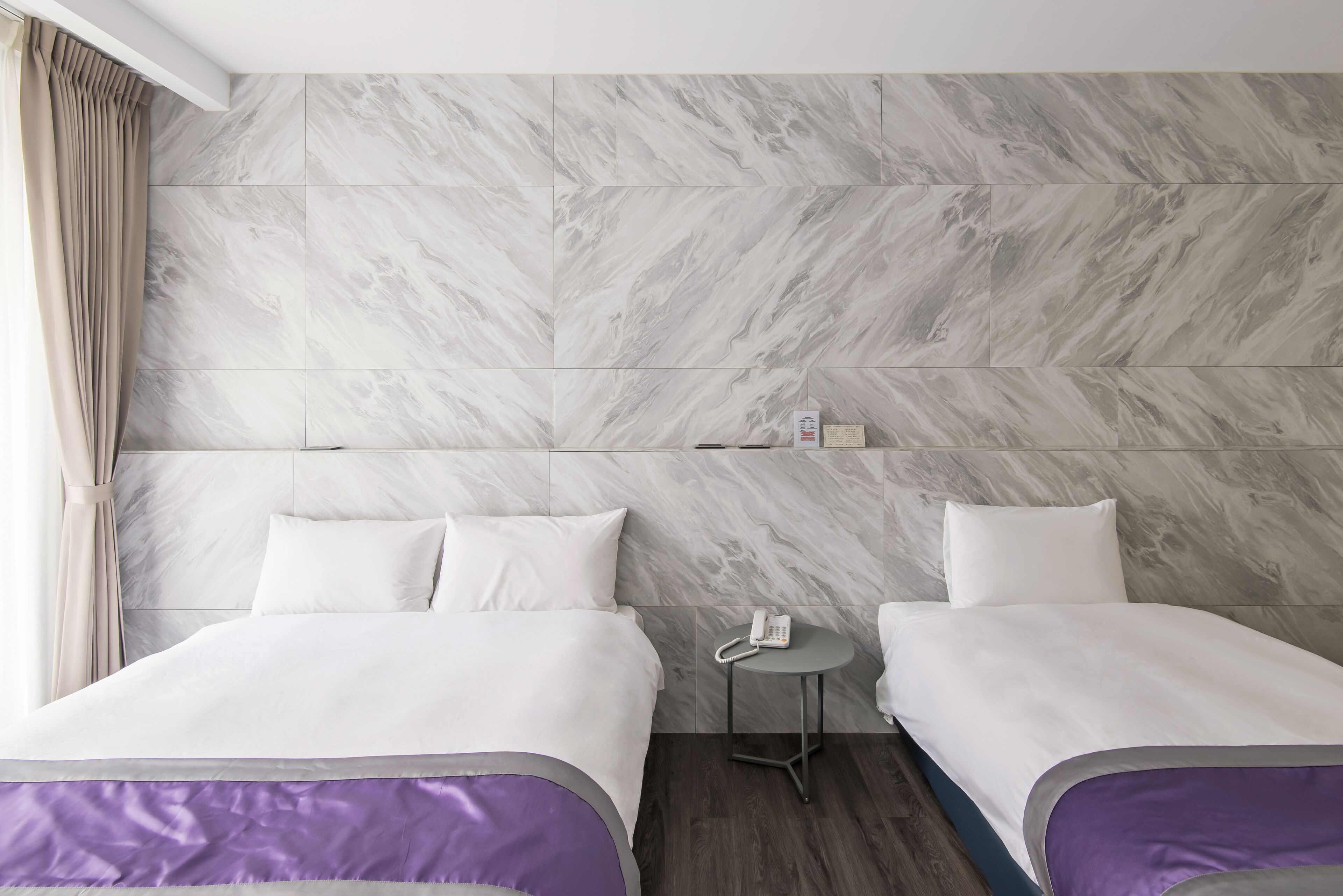 A hotel room with two beds, a small bedside table with a phone, and a marble-patterned wall.