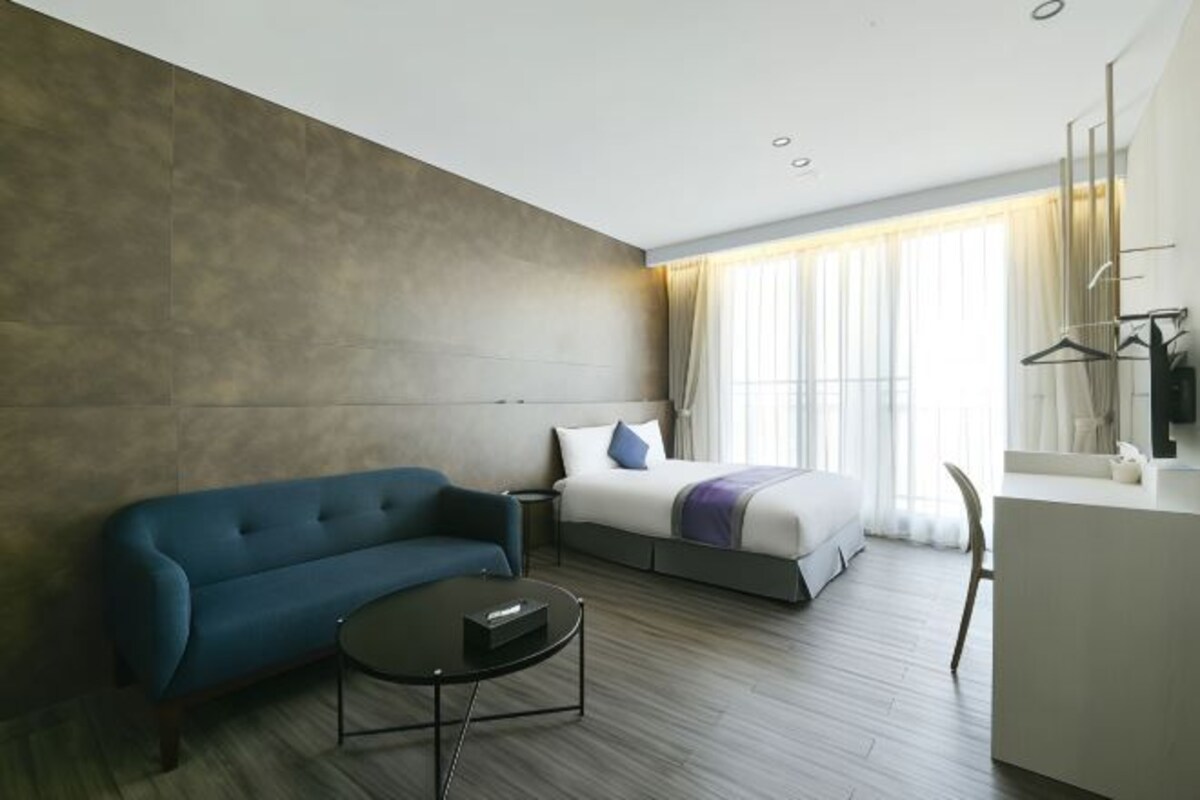 A modern hotel room with a sofa, a bed, a bedside table, and a desk.