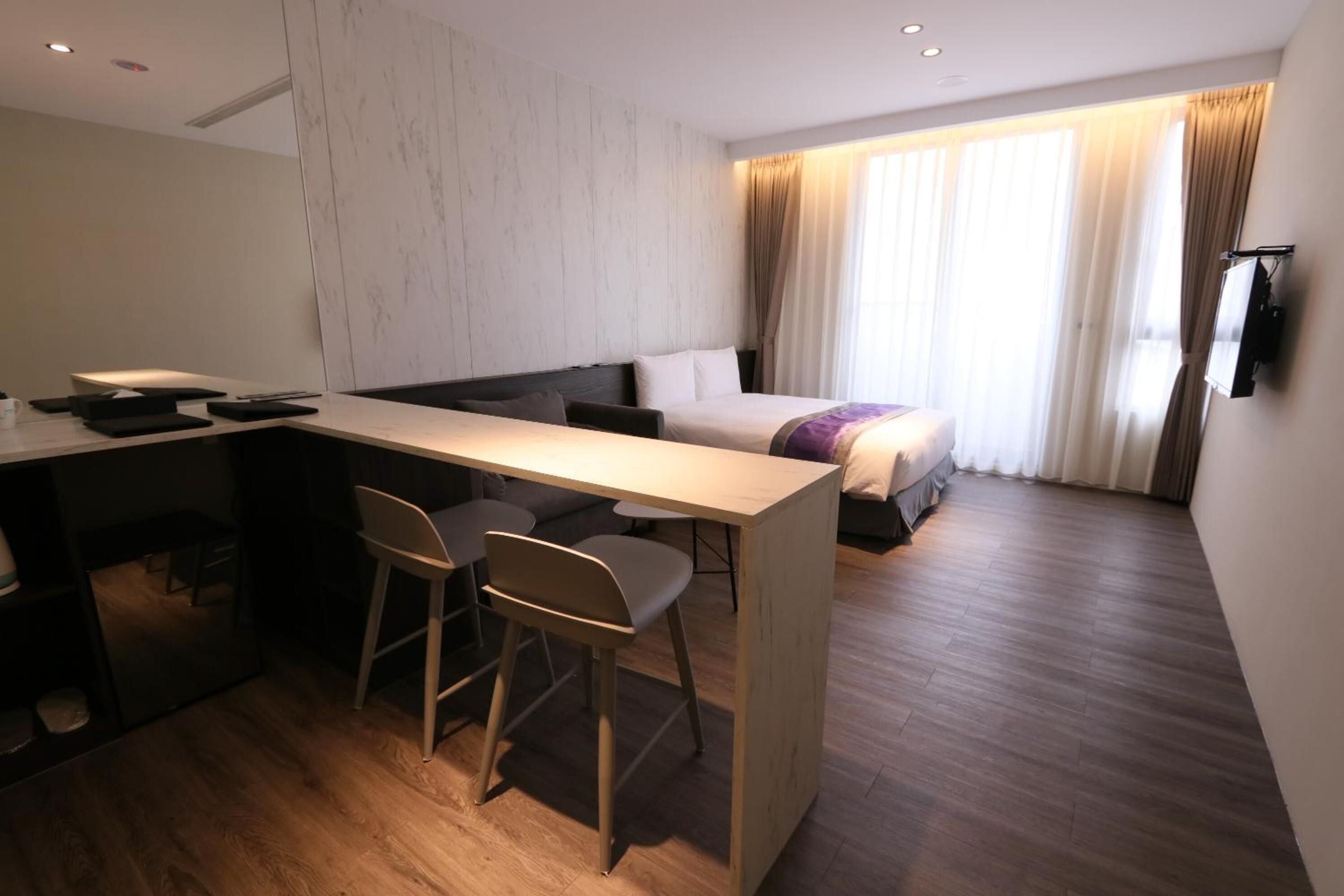 A modern hotel room with a wooden desk, two chairs, a bed with a headboard, and a TV mounted on the wall.