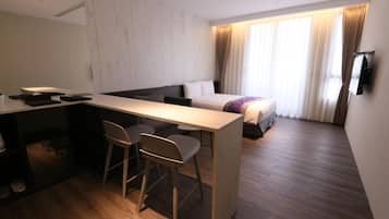 A modern hotel room with a wooden desk, two chairs, a bed with a headboard, and a TV mounted on the wall.