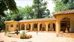A courtyard with a fountain, potted plants, and a building with arches and a tiled roof. - Holland Park (Kayunga)