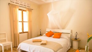 A bedroom with a bed, two bedside tables, a chair, a window with curtains, and a vase with a plant. - Holland Park (Kayunga)