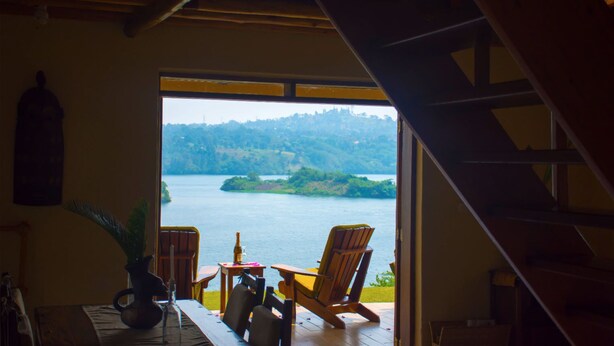 A room with a view of a lake, a wooden table with chairs, and a potted plant. - Holland Park (Kayunga)