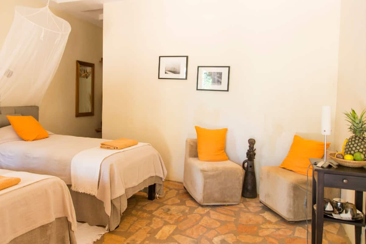 A hotel room with two beds, each with a white bedspread and orange pillows, a small armchair with an orange pillow, and a nightstand with a lamp and a fruit basket.